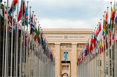 [ai] The building of the United Nations in Geneva, viewed from a pathway lined with flags of various countries, under a cloudy sky.